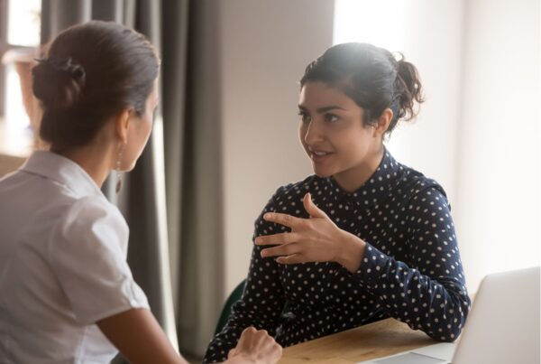 Two women sit at a desk together engaged in a conversation -- an instructional coach and a teacher.