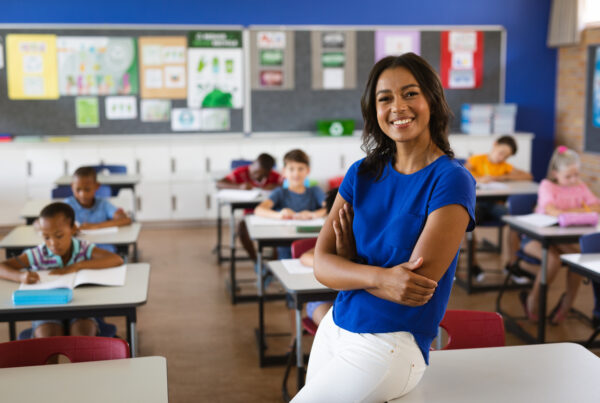 A smiling teacher in a blue top sits on the edge of a desk with her arms crossed, while elementary students work at their desks in a bright classroom behind her.